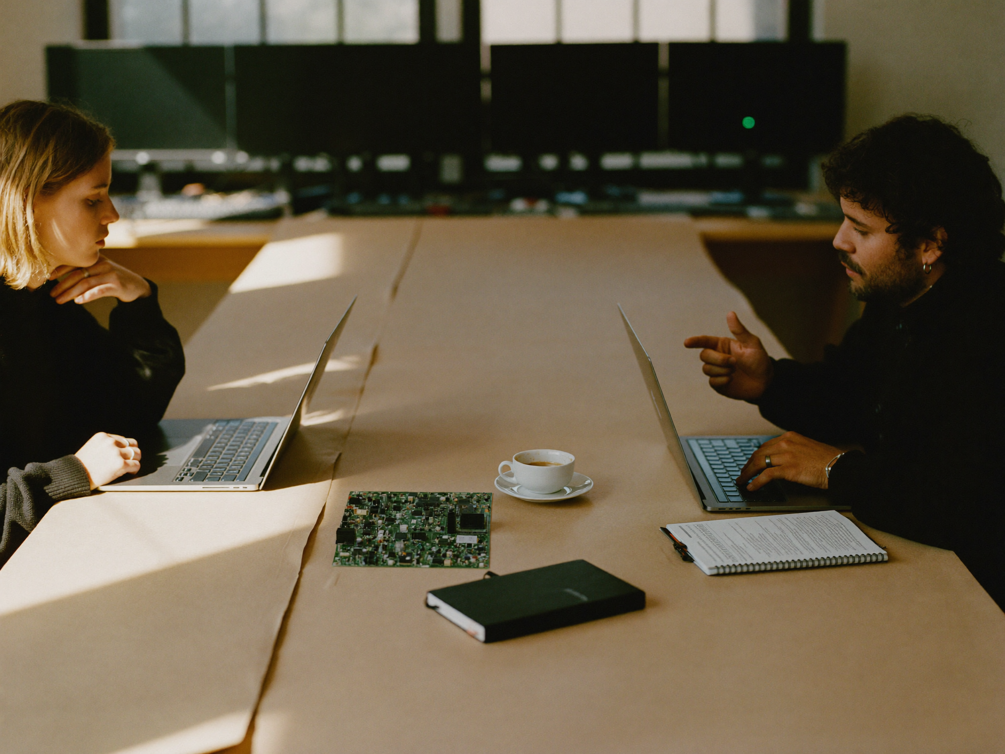 Two NewsNTech reporters at a graphite newsroom desk, mid-conversation over open laptops and a circuit board.